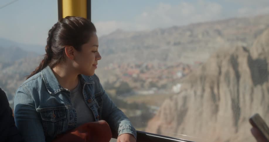 Woman rides in a cable lift up the mountain on a sunny day while someone sitting across from her holds a cell phone
