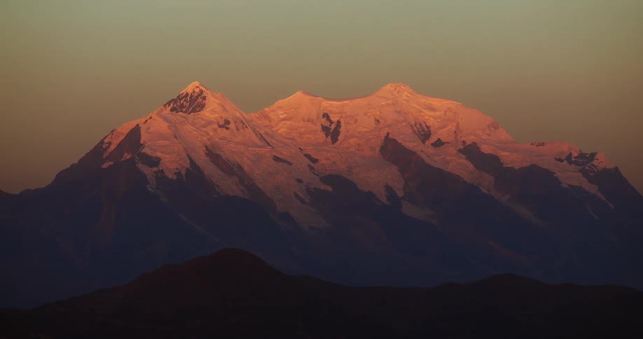 The red glow of the sun is reflecting onto the snow on a mountain ridge under a clear sky