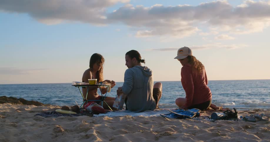 A man sitting on the beach with two women opens up some crackers and sets them on a small table