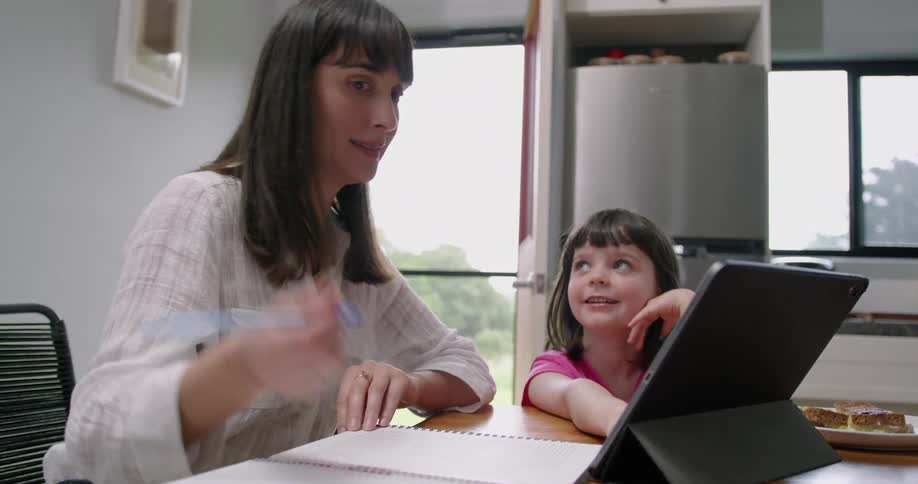 A woman is sitting at a desk in her home writing in a notebook and looking at a tablet screen as she shows her young daughter what she is doing