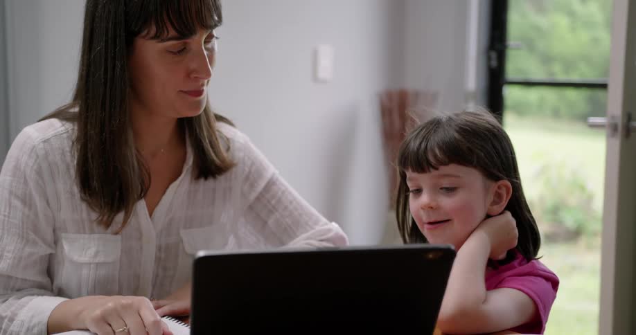 A mom points at the laptop as she talks with and teaches her daughter at home