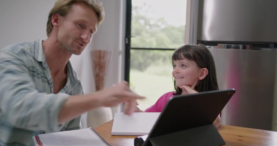 Blonde man with striped shirt sits at table helping daughter study using tablet in the kitchen