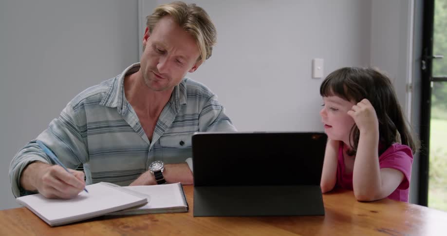 Father sitting on a table in front of a laptop at home helping his daughter with homework