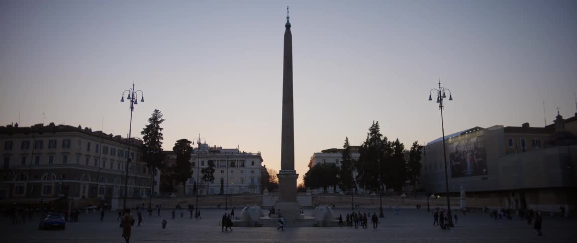 Pedestrians walking around a tall monument in a city at dusk