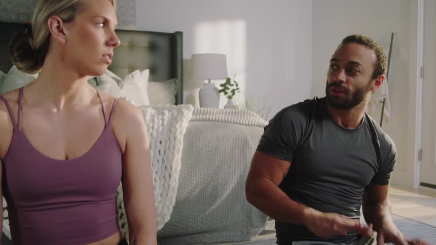 A woman and man sit on the floor in a bedroom while they have a conversation