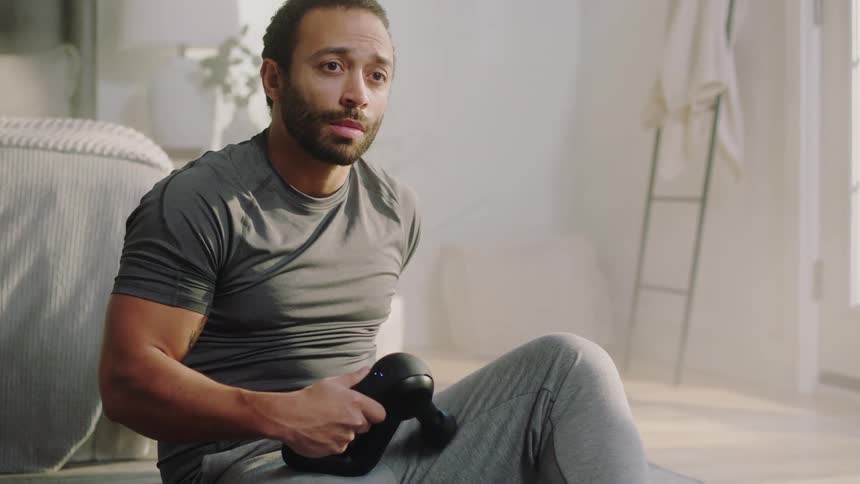 Man sitting on a bedroom floor uses a massage gun on his left leg