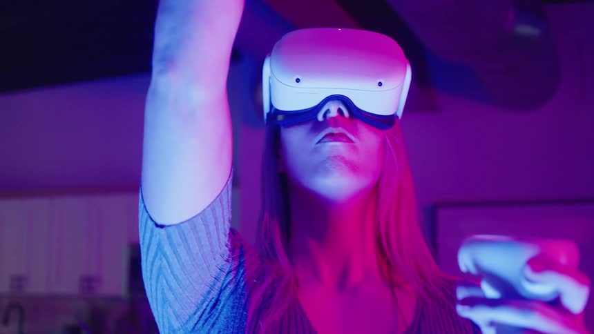 Blonde woman uses vr headset and handheld controllers while standing in room lit by colored lighting