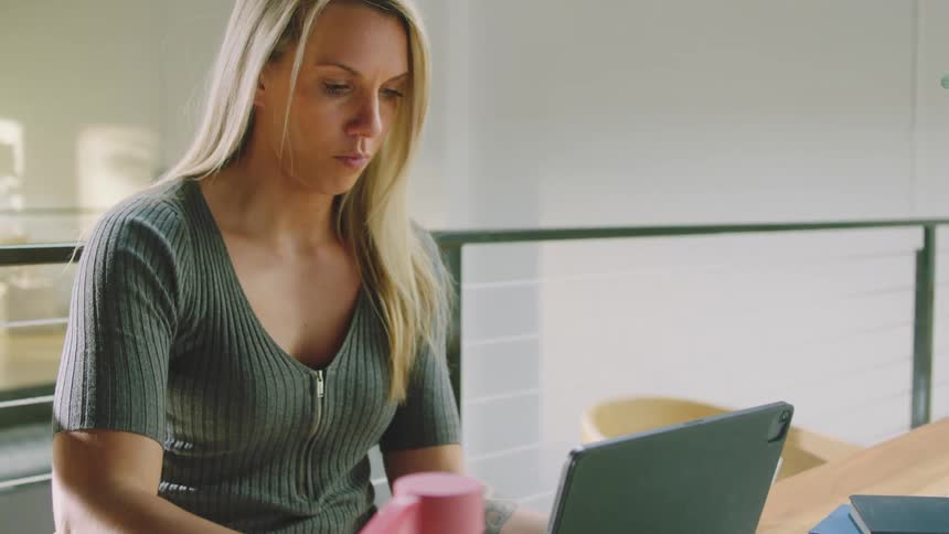 A woman drinks from her water bottle while using her tablet and writing in her notebook at a table
