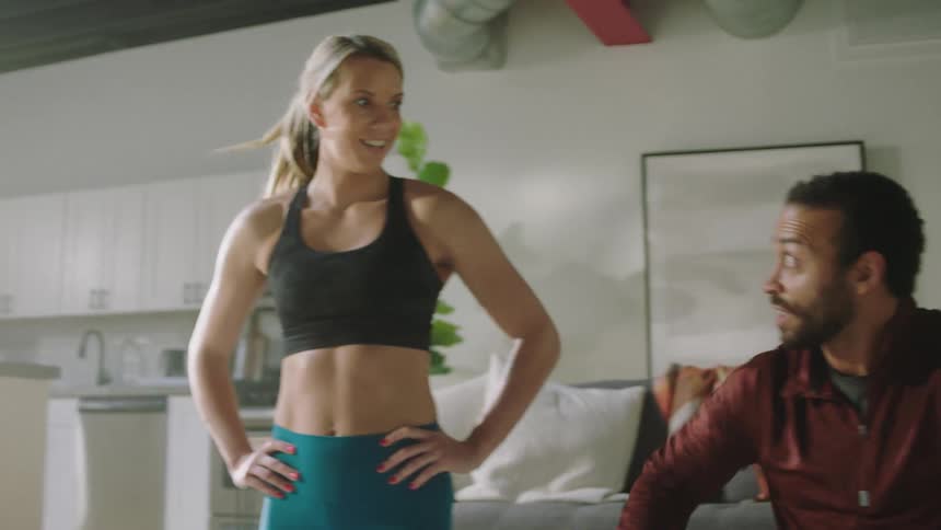 Blonde woman wearing black sports bra and teal leggings works out next to man with beard and shirt in living room at home