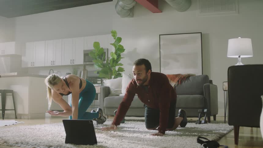 Blonde woman with black sports bra and teal leggings works out next to man with beard and shirt in living room at home