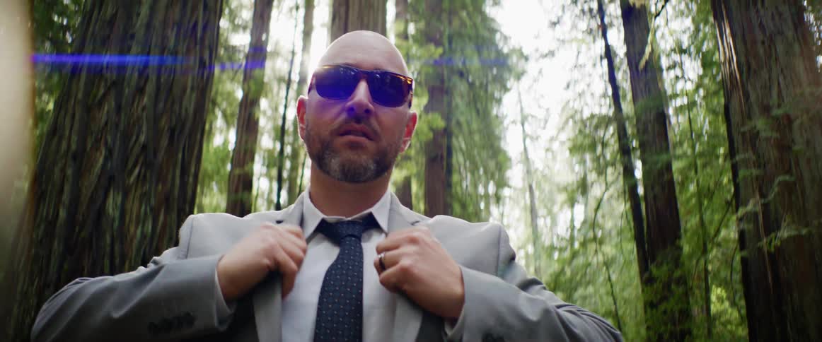 Man standing in forest among tall trees adjusts his suit jacket as sun glare glows