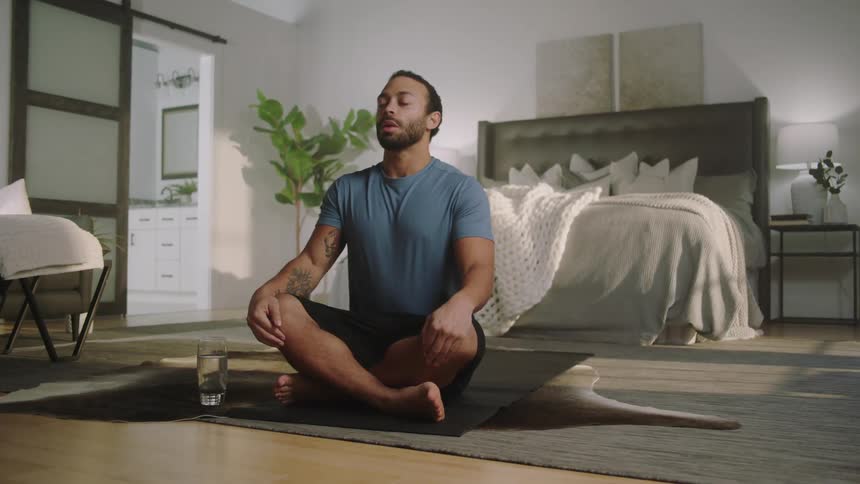 Man sets down water glass then sits cross legged on mat and starts deep breathing as he meditates