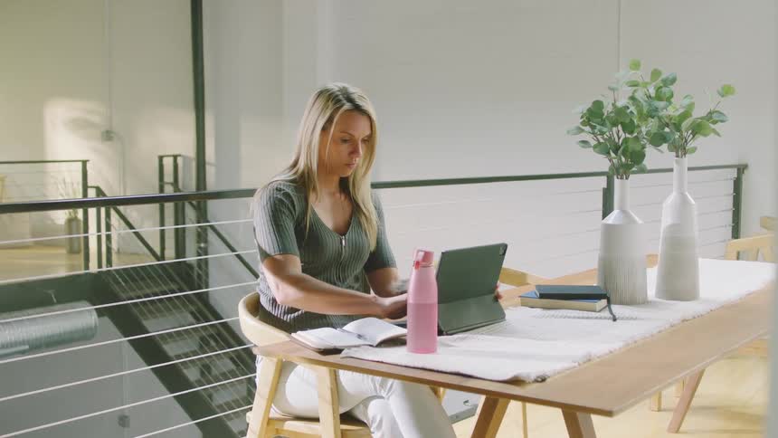 A woman sitting at a table takes a drink of her beverage before taking notes in front of her tablet computer
