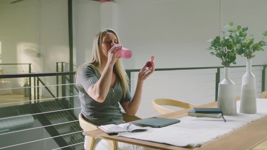 Blonde woman with green shirt and white pants drinks water from pink bottle and opens tablet at table