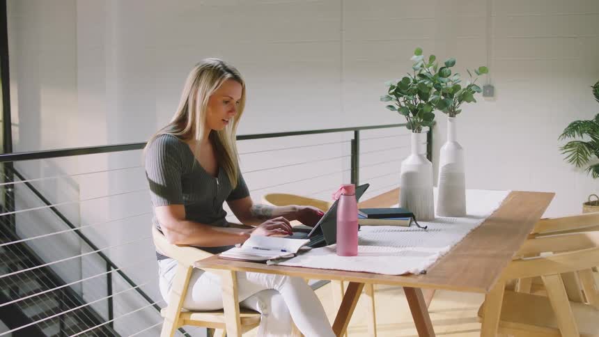 A woman is seated at a wooden table and working on a tablet