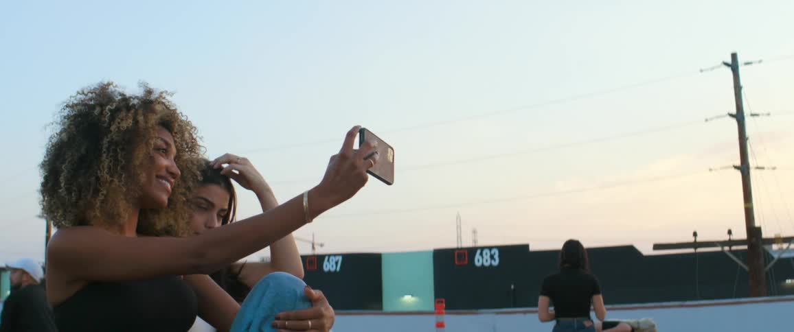 Woman holds up a cell phone to take a selfie with a woman sitting next to her as a man walks behind them and other women stand in the distance