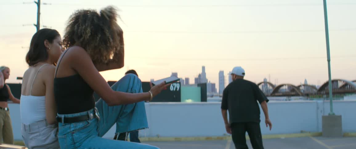 Women one holding a smartphone sit as others skate through empty parking lot with bridge and city skyline in background on foggy day