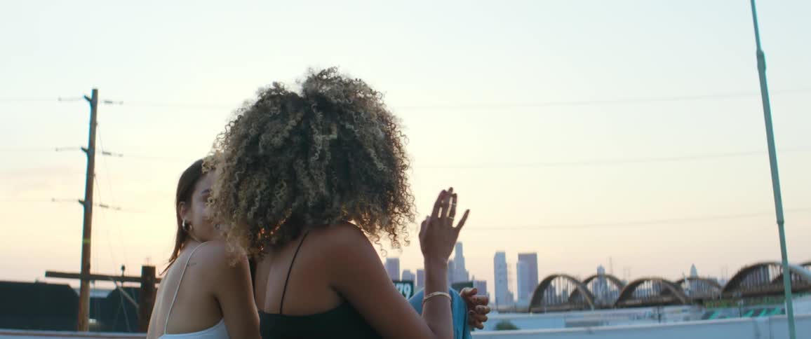 Two young female friends sittign on the roof of a building at sunset next to the city skyline