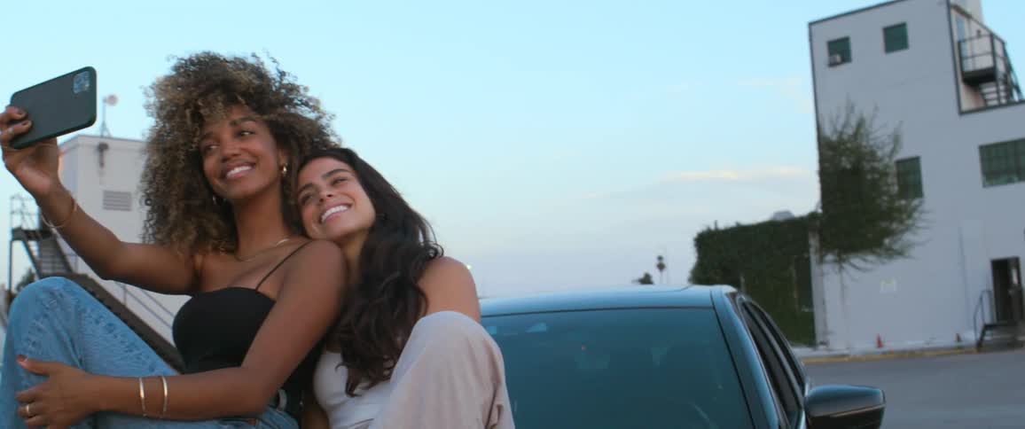 Two women sitting on the hood of a car smiling and taking selfies