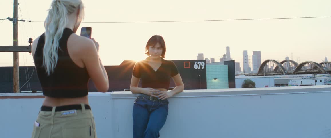 A teenager takes a photo of her friend while standing on a rooftop with views of downtown