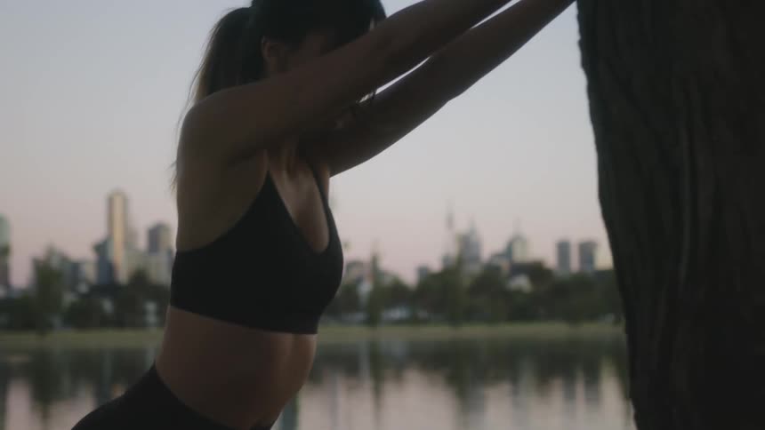 A woman pushes against a tree as she stretches out her leg muscles near a pond in a city park