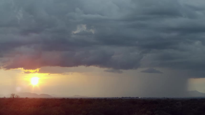 The sun glows brightly just below the clouds as lightning strikes and heavy rain falls