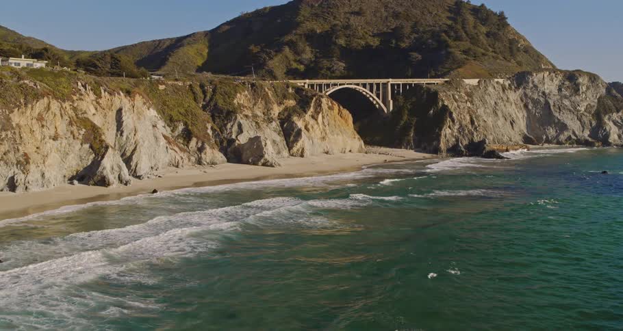 Ocean water rolls into shore near the mountains as a bird flies overhead under a blue sky