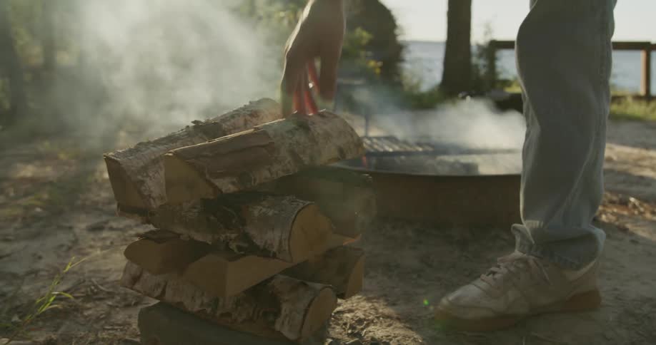Firewood is placed in a pile next to a flaming barbecue with a grill outdoors near the lake