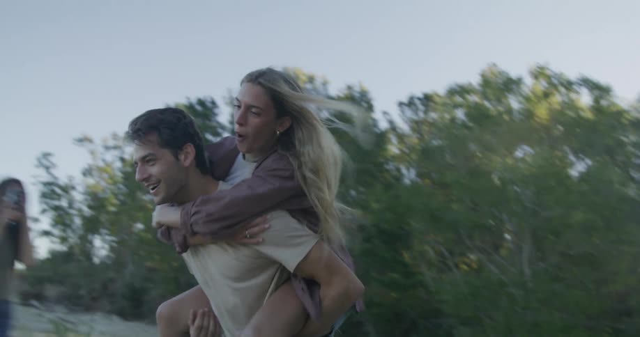 A teenage girl on a beach jumps on a boys back and he runs as she waves at friends
