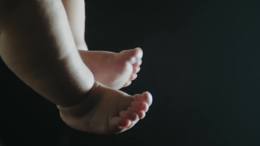 A baby rubs their feet together in front of a dark background