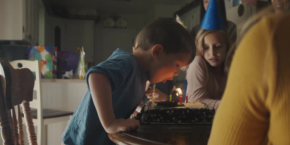 A young boy blowing out birthday candles on a cake while being cheered by friends