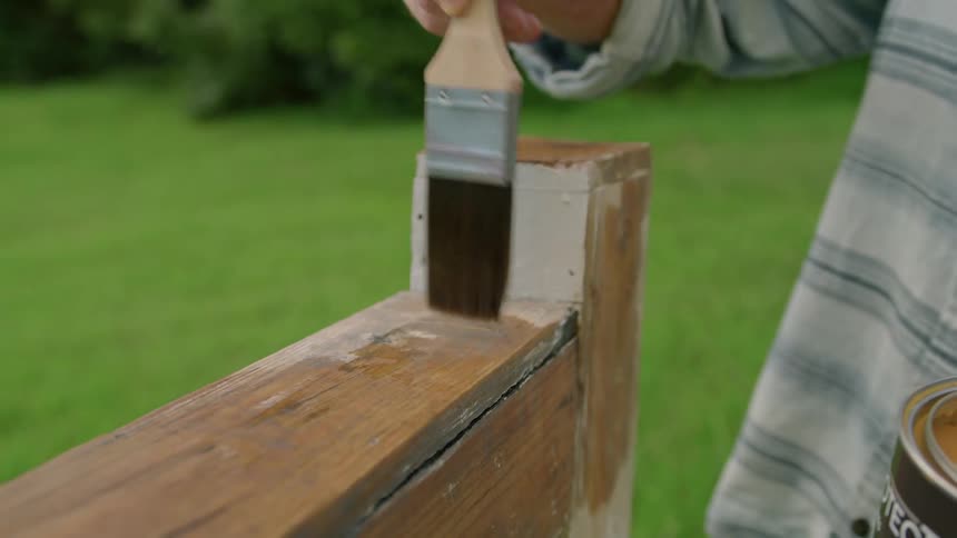A person stains a wooden object with a paintbrush outdoors