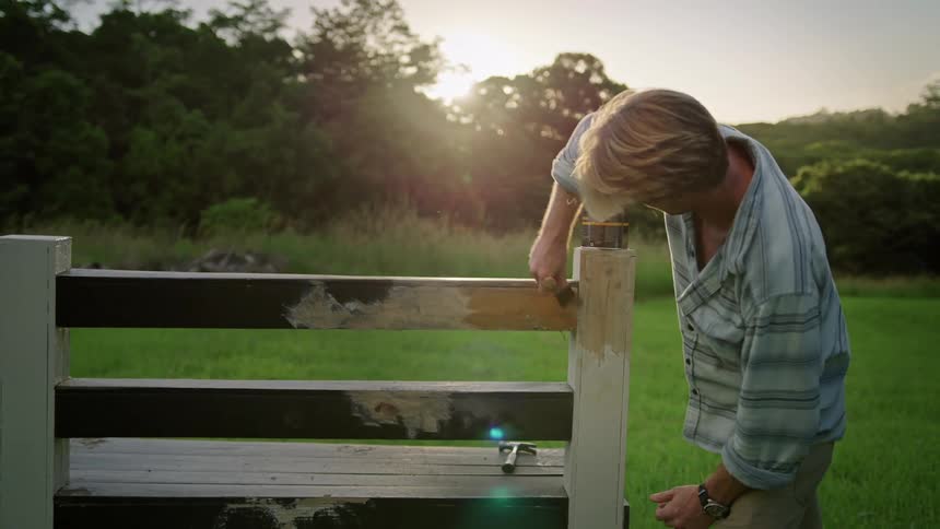 A man painting wood fencing in a rural field in the summer