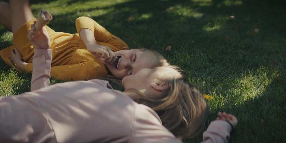 Two young sisters laughing and sharing an ice cream cone on a grassy lawn in summer