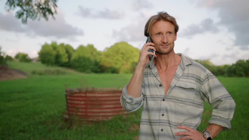 Blonde man with gray shirt and wristwatch talks on smartphone in green field in the countryside