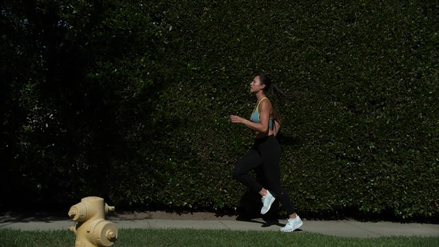 Woman jogging on sidewalk along a hedge wall in slow motion