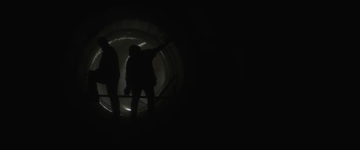 The silhouette of two men in hard hats standing in a round tunnel or pipe using a red beam flashlight