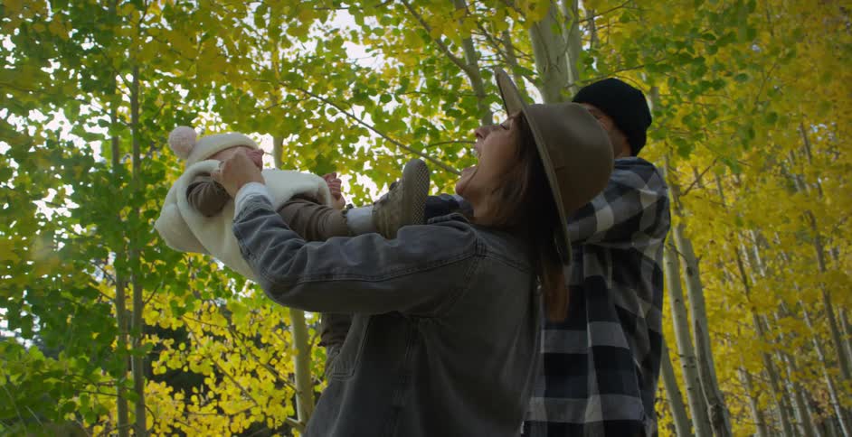 Toddler son climbs on mother wearing hat denim jacket and jeans next to father with wooly hat and gray plaid shirt in autumn forest