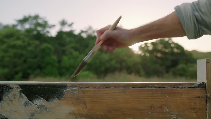 A man painting a piece of wood outside on a summer morning
