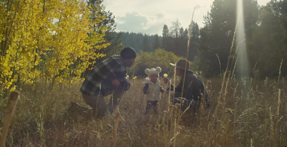 Parents smile as their daughter explores field with conifer on the horizon on sunny day