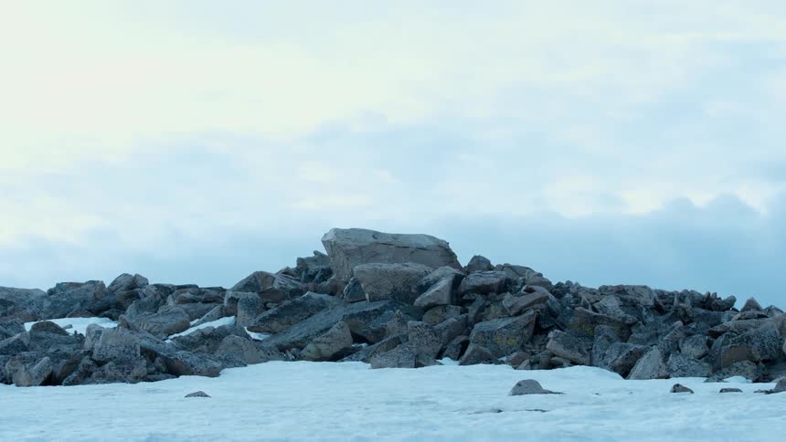 A ridge of large rocks are piled on a snow covered ground under a cloudy sky