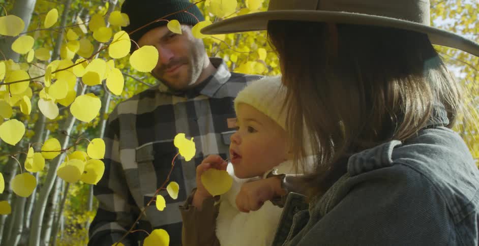 Mother with brown hat wavy hair and denim jacket holds baby son next to father with green wooly hat and gray plaid shirt in autumn forest