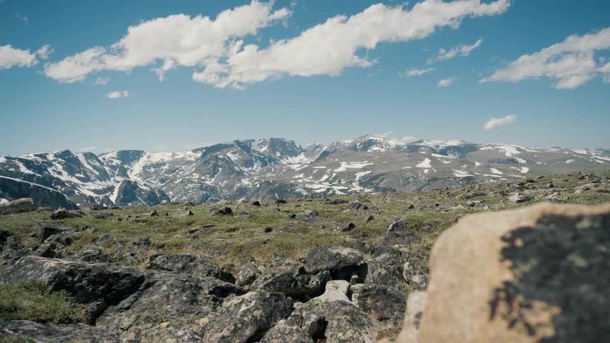 Land with large rocks and a mountain ridge with light snow under a blue sky with a few cumulus clouds