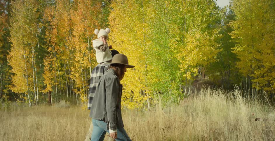 Toddler sits on a mans shoulders as they walk in a grassy field with a woman who is wearing a brimmed hat