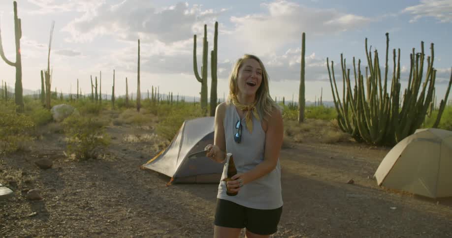 Young happy blonde woman opens a beer bottle with a knife while camping in the desert