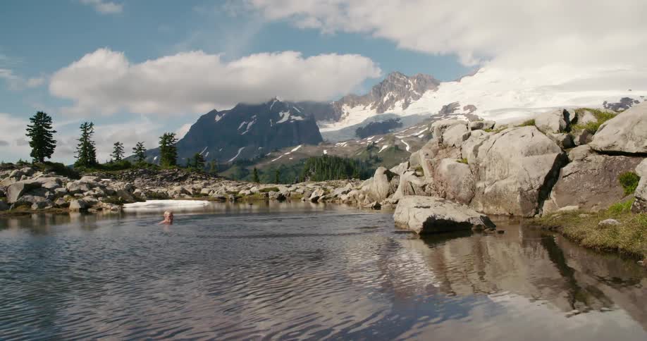 A man stands on a rock in a lake with mountains in the distance and dives in the water