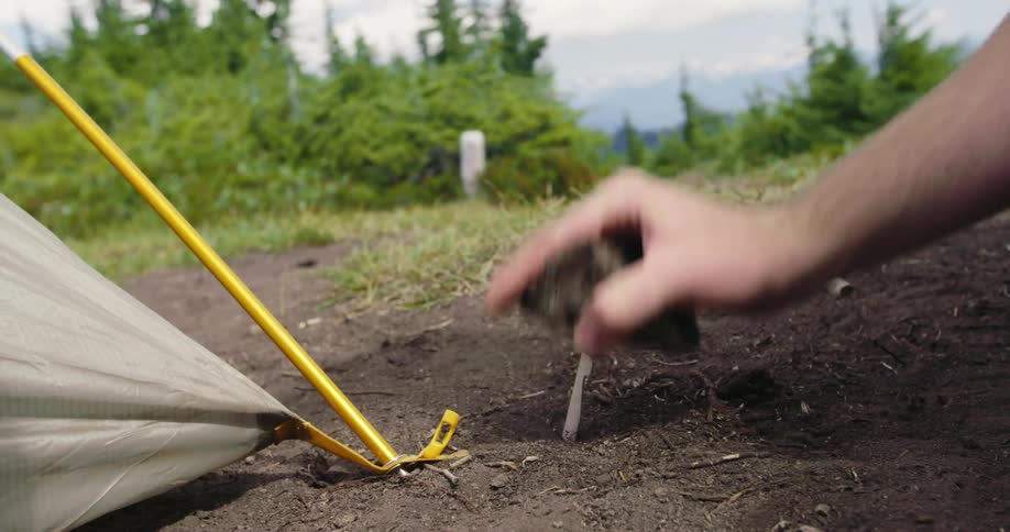 Person drives in tent stake using a stone with forest and mountains in the background