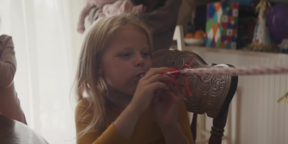 Mother holds child in the background as blonde girl plays with party favors and friends at table