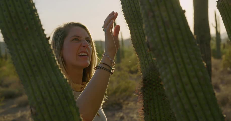 A young woman stands next to a cactus and touches the needles with her finger