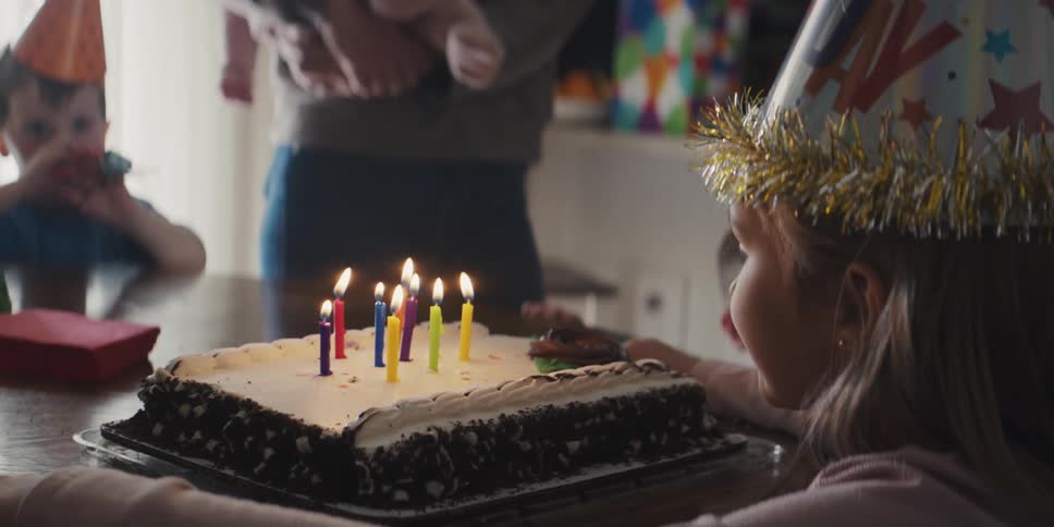 Girl with party hat blows out candles on birthday cake next to friends and parents